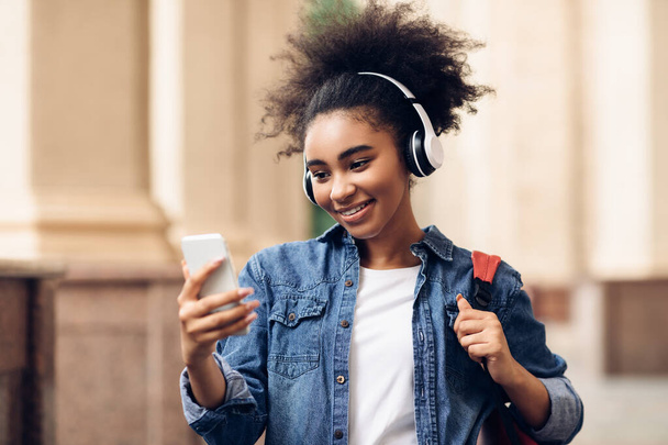 Vidám fekete diák lány használata mobiltelefon viselő fejhallgató szabadban - Fotó, kép