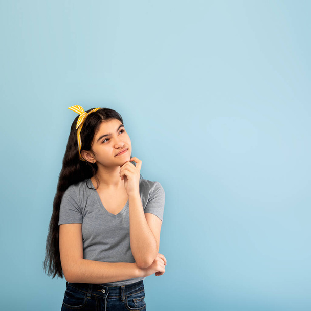 Tolles Angebot. Umfassendes indisches Teenie-Mädchen blickt in den Kopierraum und berührt ihr Kinn über blauem Studiohintergrund - Foto, Bild