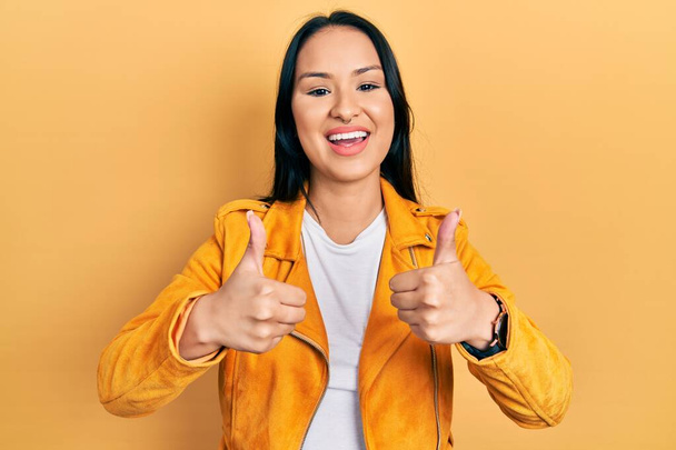 手で積極的なジェスチャーを行う黄色の革のジャケットの成功のサインを身に着けてピアス鼻を持つ美しいヒスパニック系の女性,親指を笑顔と幸せ.陽気な表情と勝者のジェスチャー.  - 写真・画像