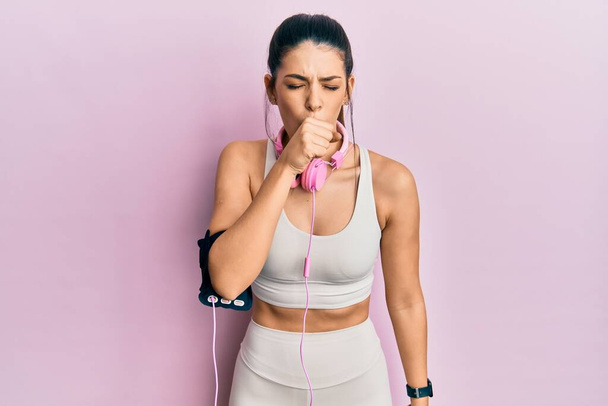 Νεαρή ισπανόφωνη γυναίκα που φοράει ρούχα γυμναστικής και χρησιμοποιεί ακουστικά που δεν αισθάνονται καλά και βήχας ως σύμπτωμα για το κρύο ή βρογχίτιδα. έννοια της υγειονομικής περίθαλψης.  - Φωτογραφία, εικόνα
