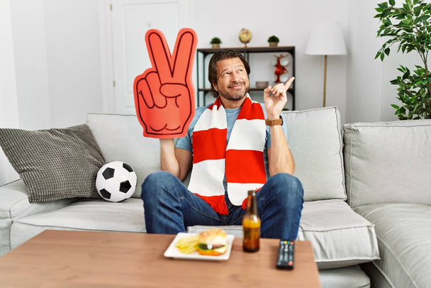 Yakışıklı orta yaşlı adam futbol holigan tezahürat oyunu evde gülümsüyor mutlu el ve parmak kenarda işaret ederek  - Fotoğraf, Görsel