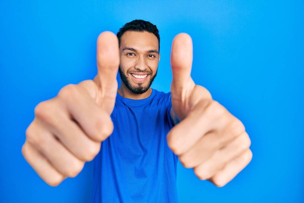髭を生やしたヒスパニック系の男性は、手で積極的なジェスチャーを行うことを承認カジュアルブルーのTシャツを着て、親指を上げて笑顔と成功のために幸せ。勝者のジェスチャー.  - 写真・画像
