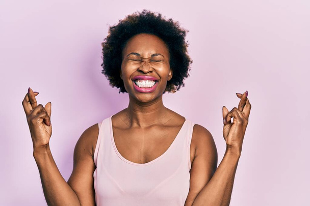 Young african american woman wearing casual sleeveless t shirt gesturing finger crossed smiling with hope and eyes closed. luck and superstitious concept.  - Photo, Image