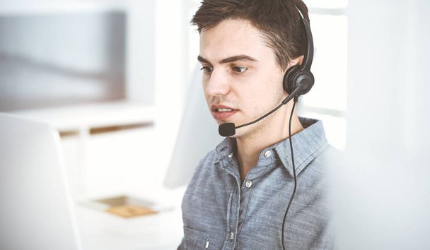 Casual dressed young man using headset and computer while talking with customers online in sunny office. Call center, business concept - Valokuva, kuva
