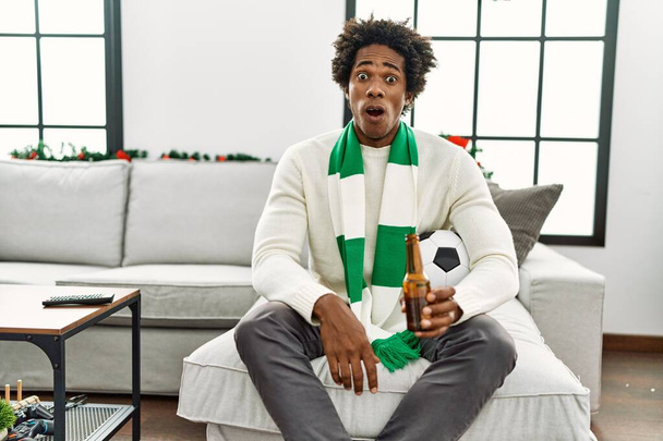 Νεαρός Αφροαμερικάνος ποδοσφαιριστής χούλιγκαν πίνοντας μπύρα ζητωκραυγάζοντας παιχνίδι φοβισμένος και έκπληκτος με ανοιχτό το στόμα για έκπληξη, δυσπιστία πρόσωπο  - Φωτογραφία, εικόνα