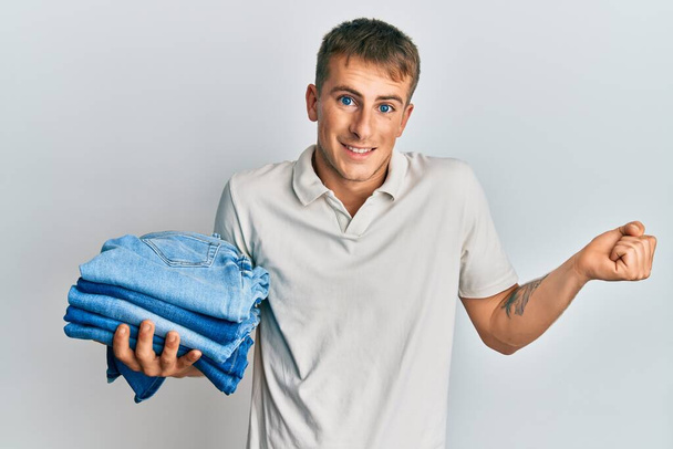 Young caucasian man holding stack of folded jeans screaming proud, celebrating victory and success very excited with raised arm  - Foto, imagen