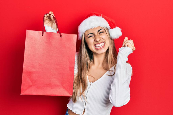 Mooie Spaanse vrouw draagt kerstmuts en houdt boodschappentas schreeuwend trots, viert overwinning en succes zeer enthousiast met opgeheven arm  - Foto, afbeelding