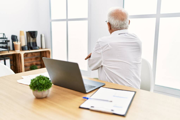 Homme âgé travaillant au bureau à l'aide d'un ordinateur portable debout vers l'arrière regardant loin avec les bras croisés  - Photo, image