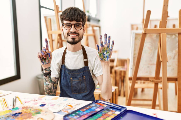 Spanyol férfi szakállal a művészeti stúdióban, mutatva és mutatva a hatodik ujjal, miközben magabiztosan és boldogan mosolyog..  - Fotó, kép