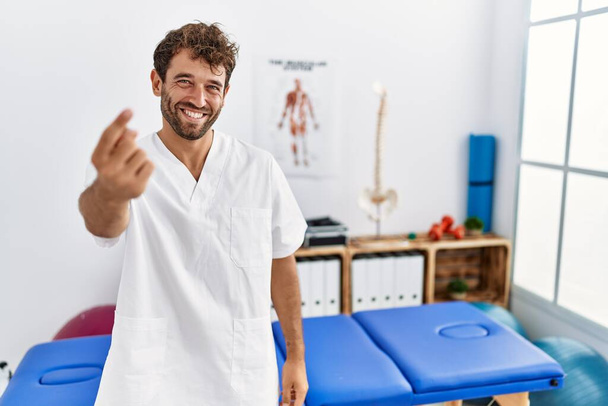 Junge gut aussehende Physiotherapeutin, die in der Schmerzklinik arbeitet, winkt mit einer Handbewegung, die sie einlädt, glücklich und lächelnd zu sein  - Foto, Bild