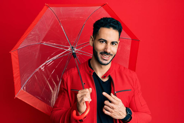Young hispanic man holding umbrella smiling happy pointing with hand and finger  - Photo, Image