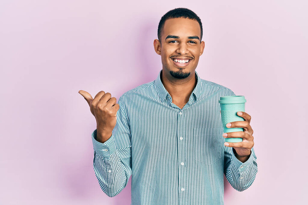 Joven afroamericano bebiendo una taza de café para llevar apuntando con el pulgar hacia un lado sonriendo feliz con la boca abierta  - Foto, imagen