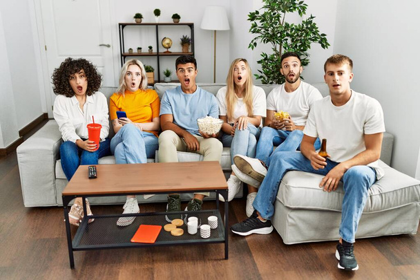 Grupo de pessoas comendo lanche sentado no sofá em casa assustado e surpreso com a boca aberta para surpresa, rosto descrença  - Foto, Imagem