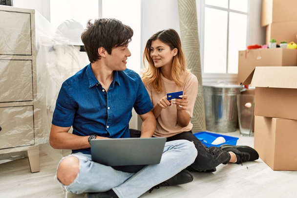 Jeune couple caucasien utilisant ordinateur portable et carte de crédit assis sur le sol à la nouvelle maison. - Photo, image