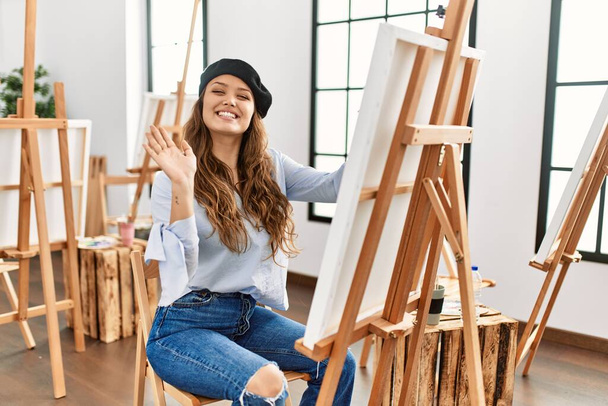 Junge hispanische Künstlerin malt auf Leinwand im Kunstatelier und verzichtet auf fröhliches Hallo und lächelnde freundliche Willkommensgeste  - Foto, Bild