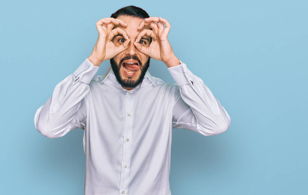 Young man with beard wearing business shirt doing ok gesture like binoculars sticking tongue out, eyes looking through fingers. crazy expression.  - Photo, image