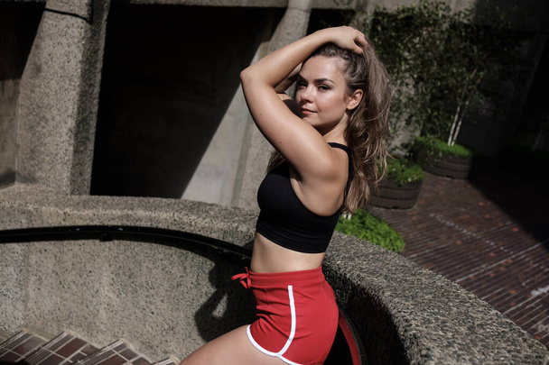 Joven mujer de fitness posando junto a una barandilla. Ella está usando un top negro y pantalones cortos deportivos rojos. - Foto, imagen
