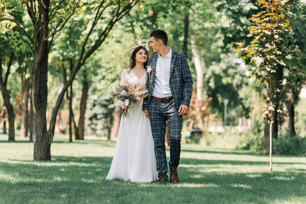 Wedding, Happy young groom and beautiful tender bride in a wedding dress with a bouquet, Bride and groom hold hands and walk on a green field, wedding day, wedding ceremony in boho style - Zdjęcie, obraz