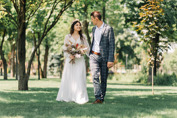 Wedding, Happy young groom and beautiful tender bride in a wedding dress with a bouquet, Bride and groom hold hands and walk on a green field, wedding day, wedding ceremony in boho style - Foto, Imagen