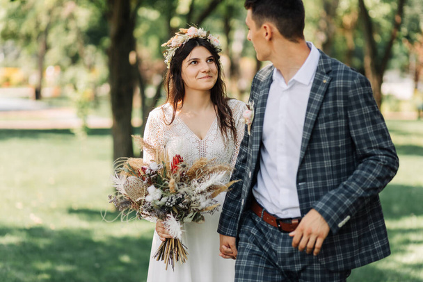 Wedding, Happy young groom and beautiful tender bride in a wedding dress with a bouquet, Bride and groom hold hands and walk on a green field, wedding day, wedding ceremony in boho style - Fotó, kép