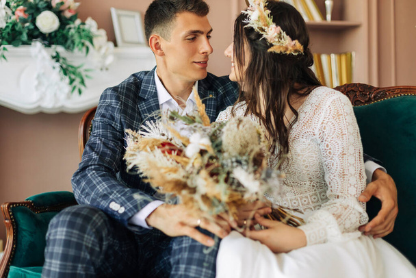Happy young couple, stylish groom in a suit and a young bride in a wedding lace dress with a bouquet of flowers, sit together, hug, in the studio on the sofa, wedding in boho style - Foto, Imagen