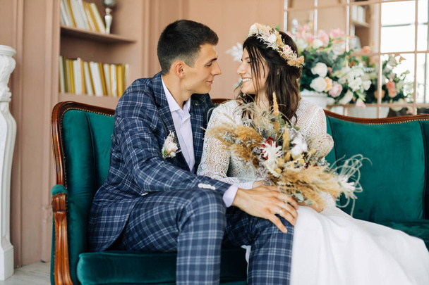 Happy young couple, stylish groom in a suit and a young bride in a wedding lace dress with a bouquet of flowers, sit together, hug, in the studio on the sofa, wedding in boho style - Photo, Image