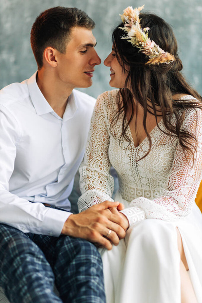 Young groom in a gray suit and a beautiful smiling bride in a white lace dress, Wedding couple sit in an armchair on a gray wall background, Wedding portrait of a couple in love - Foto, Imagem