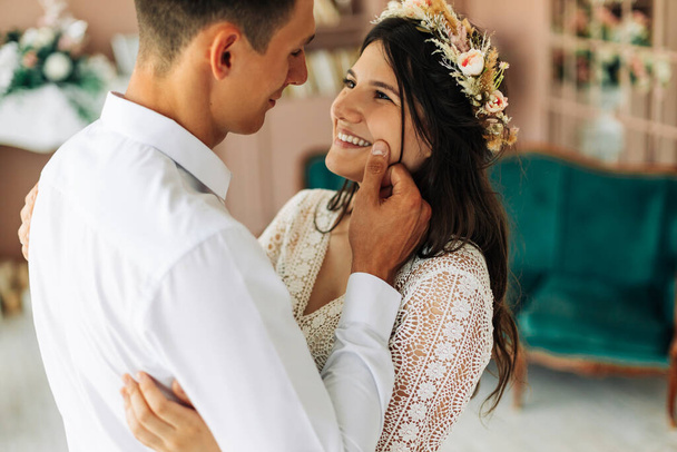 Happy young couple, stylish groom in a suit and a young bride in a wedding lace dress with a bouquet of flowers, sit together, hug, in the studio on the sofa, wedding in boho style - Φωτογραφία, εικόνα
