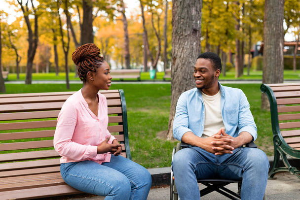 Cheerful disabled black guy in wheelchair having conversation with his girlfriend at park in autumn - Foto, immagini