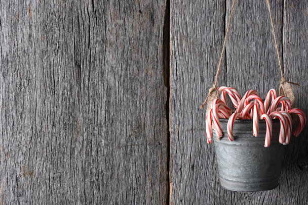 Christmas Candy Canes: A bucket full of the peppermint holiday treat hanging from twine against a rustic wood wall with copy space. - Photo, image