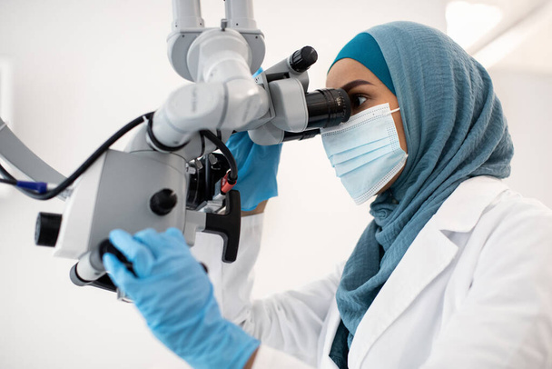 Ισλαμική οδοντίατρος γυναίκα φορώντας προστατευτική ιατρική μάσκα χρησιμοποιώντας οδοντιατρικό μικροσκόπιο στην κλινική - Φωτογραφία, εικόνα