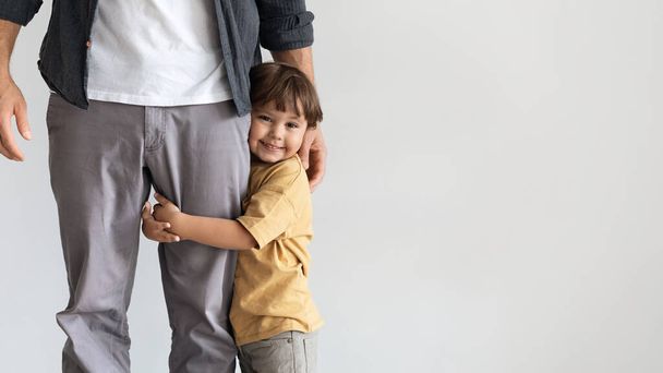 Happy little boy embracing his daddy, smiling to camera, feeling loved and safe, posing over grey wall with empty space - Photo, Image