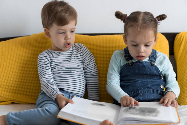 Двое детей дошкольного возраста читают на жёлтой кровати большую интересную книгу сказок. Братья и сёстры, читающие близнецов, веселятся, счастливые дети дома на карантине. Концепция дружбы, семьи, образования - Фото, изображение
