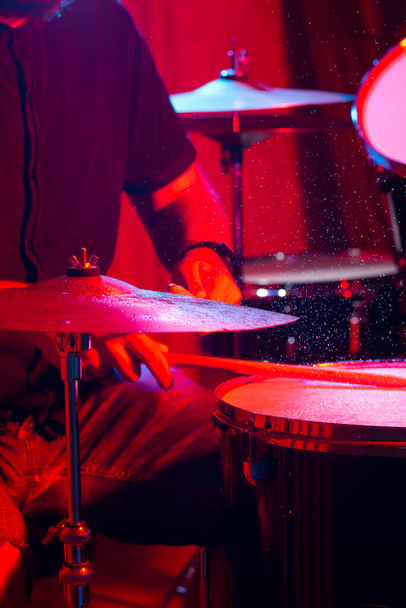 uomo suona strumento musicale a percussione con bastoni con spruzzi d'acqua da vicino su fondo nero, suonare al tamburo, illuminazione rossa e blu sul palco. - Foto, immagini
