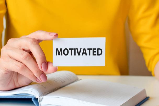 motivated is written on a white business card. A woman's hand holds a white paper card against the background of a yellow dress and a white notebook. Business and advertising concept. - Valokuva, kuva