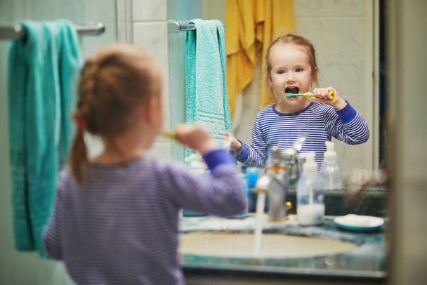 Happy toddler girl in pyjamas brushing her teeth in bathroom in the morning or before going to sleep. Dental hygiene for small kids - Foto, imagen