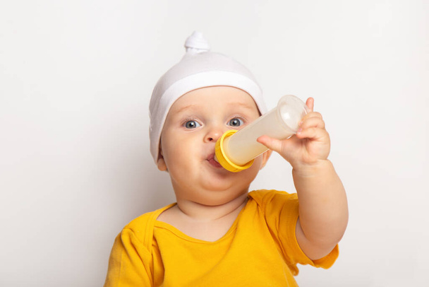portrait of a cute happy caucasian baby boy dressed in a yellow t-shirt and a hat on white background. child drinks milk from a bottle with copyspace. nutrition and healthcare concept - Foto, immagini