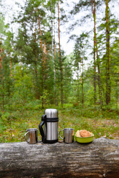 松林の中のピクニック。森を背景に古い丸太の上に魔法瓶や金属製のマグカップが立っている。垂直フレームと選択フォーカス. - 写真・画像