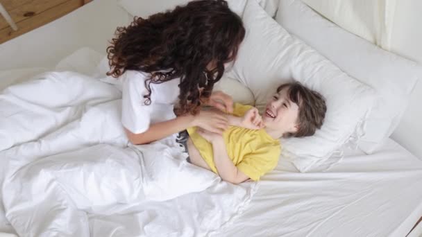Happy mom tickling laughing boy lying in bed. Caring cheerful mother wake up kid on morning weekend - Footage, Video
