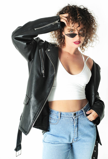 Stylish curly hair woman in sunglasses and leather jacket posing on white background. - Фото, изображение