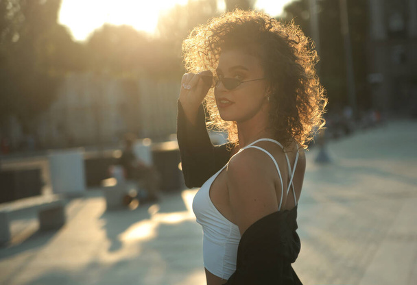 Stylish curly hair woman in sunglasses and leather jacket posing outdoors at sunset. - Zdjęcie, obraz
