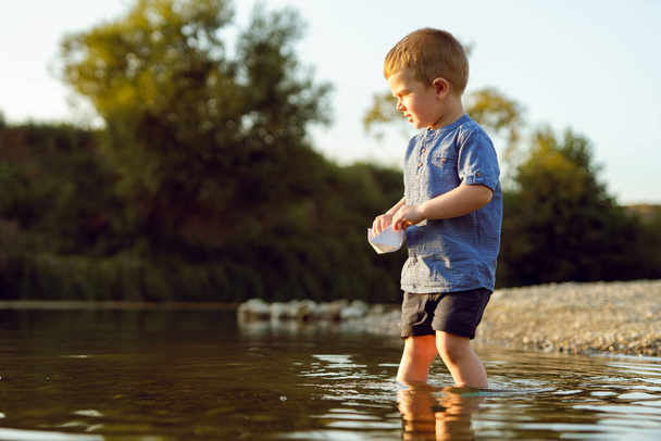Pequeno caucasiano menino loiro cabelo três anos de idade sozinho no rio ou lago em verão ensolarado ou outono à noite brincando com papel barco infância tranquilidade conceito copiar espaço - Foto, Imagem