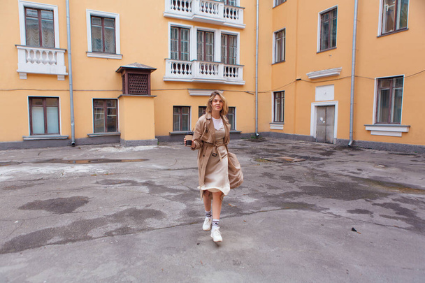Young smiling millennial woman with wild hair dressed in an autumn coat walking with a cup of coffee to go near the old building. - Valokuva, kuva