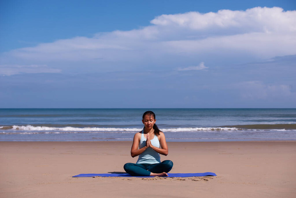 люди, фитнес, спорт и здоровый образ жизни концепции - молодая азиатская женщина делает медитацию на тропическом пляже с голубым небом фоне - Фото, изображение