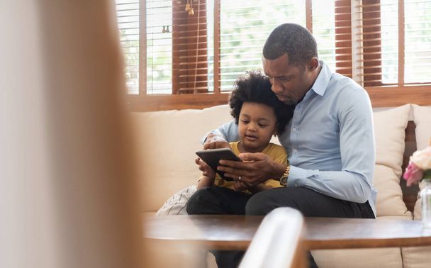 Happy Black African American Brazilian father and little boy sitting on sofa using digital tablet in living room at home together. - 写真・画像