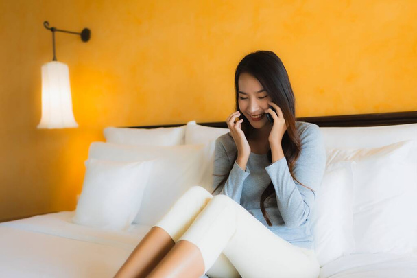 Yatağın üzerinde cep telefonu ve yatak odasının içinde kahve fincanı kullanan güzel Asyalı kadın portresi. - Fotoğraf, Görsel