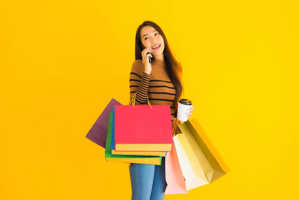 Schöne junge asiatische Frau verwenden Smartphone oder Handy mit Kaffeetasse und farbiger Einkaufstasche auf gelbem Hintergrund - Foto, Bild