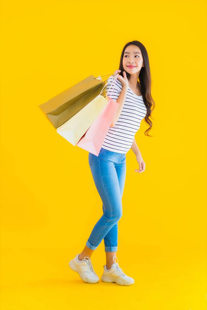 黄色の隔離された背景にカラフルなショッピングバッグを持つ肖像画の美しい若いアジアの女性 - 写真・画像
