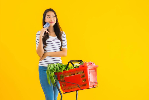 Retrato bonito jovem asiático mulher compras carrinho de supermercado com cartão de crédito no fundo isolado amarelo
 - Foto, Imagem