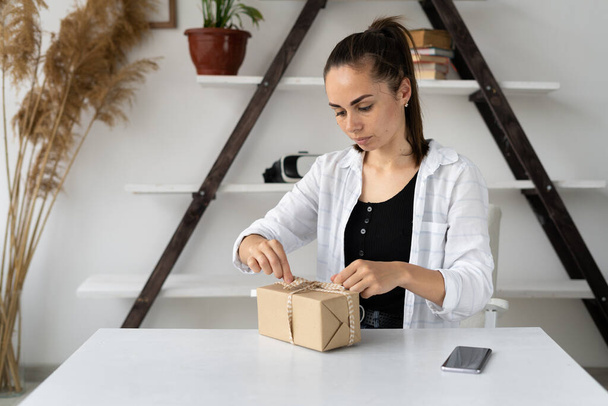 Young caucasian woman is sitting at the table and packing a box. Small business concept at home preparing goods for shipment to the buyer. Copy space. - Fotó, kép